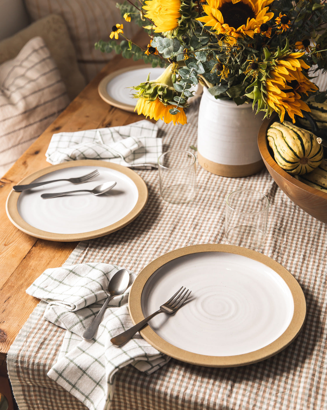 Dining table setting with plates, cutlery, and a vase of sunflowers on a checkered tablecloth.