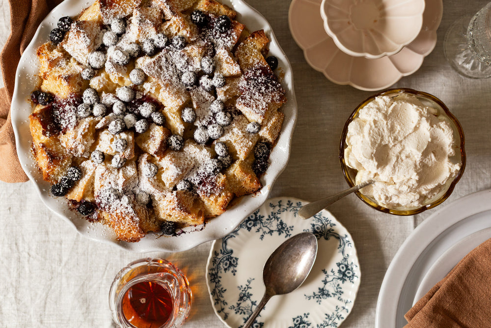 Baked pudding with berries and cream on a table with plates and a spoon.