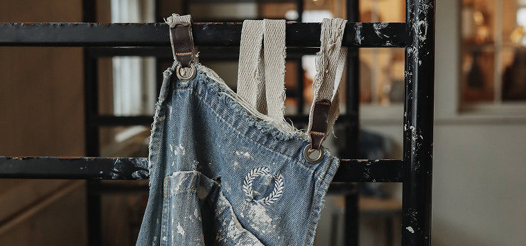 Worn denim overalls with suspenders hanging on a metal rack.